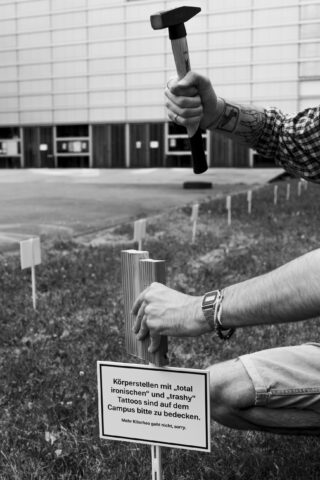 Vor einem modernen Gebäude rammt eine Person einen Holzpfahl in den Boden und hält ein Schild mit deutscher Aufschrift über Tätowierungen auf dem Campus hoch.