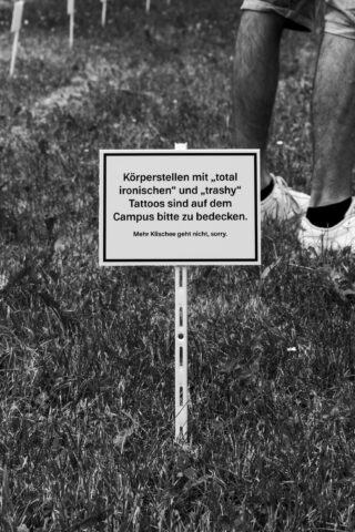 Auf einer Rasenfläche ruft ein Schild in deutscher Sprache dazu auf, „total ironische“ und „trashige“ Tattoos auf dem Campus abzudecken. In der Nähe stehen Menschen, von denen nur ihre Beine zu sehen sind.