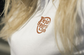 Nahaufnahme eines weißen Trikots mit dem orangefarbenen Aufdruck „Tejo Sport Club“. Langes, blondes Haar bedeckt das Trikot teilweise.