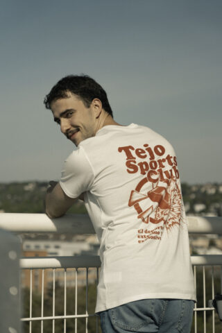 Ein Mann lehnt im Freien mit abgewandtem Blick an einem weißen Geländer. Er trägt ein weißes T-Shirt mit der roten Aufschrift „Tejo Sports Club“ auf dem Rücken. Im Hintergrund sind Bäume und Gebäude zu sehen.