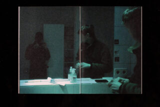 In einem Badezimmerspiegel spiegeln sich zwei Personen. Eine hält eine Kamera in der Hand, die andere steht mit Pflegeprodukten und einer Flasche Handdesinfektionsmittel auf der Ablage am Waschbecken.