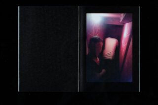 Ein schwach beleuchteter Flur mit einer Person im Vordergrund, die der Kamera zugewandt ist, und einer anderen Person, die am Ende des Korridors zur Wand blickt.