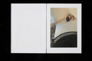 Ein offenes Buch zeigt das Foto einer Person, die zusammengerollt auf einer nackten Matratze neben einer Wand liegt. Im Vordergrund ist ein großer Ventilator teilweise sichtbar. Die gegenüberliegende Seite ist leer.
