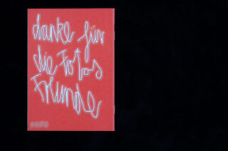 Ein rotes Büchlein mit handgeschriebenem weißem Text auf Deutsch mit der Aufschrift „Danke für die Fotos Freunde“ und „KOKO“ in der unteren linken Ecke vor schwarzem Hintergrund.