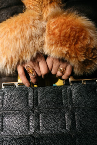 Nahaufnahme von Händen mit goldenen Ringen und einem pelzbesetzten Mantel, die eine strukturierte schwarze Handtasche halten.
