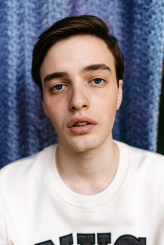 Ein junger Mann mit kurzen braunen Haaren und blauen Augen trägt ein weißes Sweatshirt und blickt mit neutralem Gesichtsausdruck direkt in die Kamera. Im Hintergrund hängt ein violetter Stoff mit Struktur.