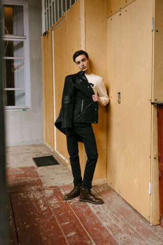 Eine Person steht in einem Gebäude vor einer Sperrholzwand, hält eine schwarze Lederjacke über der Schulter und trägt dunkle Hosen und braune Stiefel.