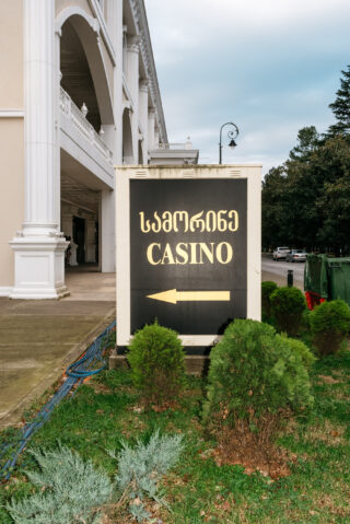 Ein Schild mit dem Wort „CASINO“ und Text in einer anderen Sprache weist nach links und ist vor einem Gebäude mit Säulen und Grünanlagen angebracht.