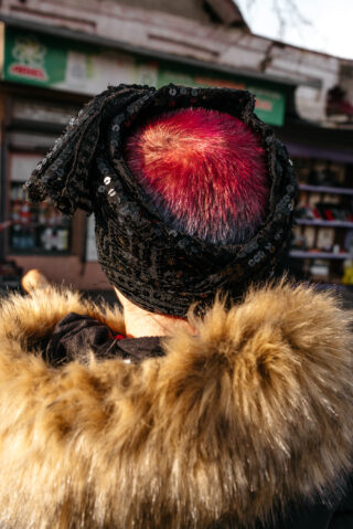 Eine Person mit kurzen, leuchtend roten Haaren trägt einen schwarzen Paillettenschal und einen pelzbesetzten Mantel, von hinten fotografiert auf einer Stadtstraße.