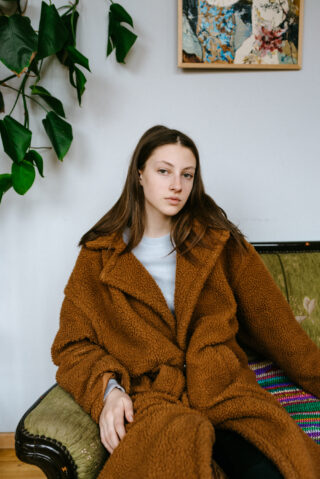 Eine junge Frau mit langen braunen Haaren sitzt auf einem grünen Sofa und trägt einen braunen, flauschigen Mantel. Im Hintergrund sind eine Pflanze und ein gerahmtes Kunstwerk zu sehen.