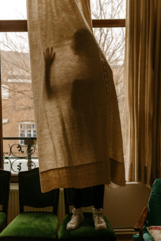 Eine Person steht auf einem grünen Stuhl hinter einem beigen Vorhang. Ihre Silhouette und Hand sind durch den Stoff in einem Raum mit großen Fenstern sichtbar.