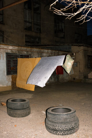 Wäsche hängt auf einer Wäscheleine zwischen zwei alten Reifen in einem schwach beleuchteten, städtischen Innenhof mit verwitterten Gebäuden im Hintergrund.