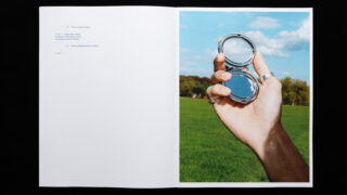 Eine Hand hält einen offenen Taschenspiegel vor einer Wiese und blauem Himmel, in dem sich ein Teil der Hand und des Himmels spiegelt.