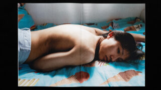 Eine Person liegt ohne Hemd auf dem Bauch auf einem Bett mit blauen Laken, auf denen Eistüten gemustert sind, und blickt in die Kamera.