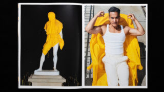 In einer aufgeschlagenen Zeitschrift ist links das Foto einer in ein gelbes Tuch gehüllten Statue zu sehen, und rechts ein Mann, der auf einem gelben Mantel liegend die Arme streckt.