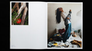 Offenes Buch mit zwei Seiten: Auf der linken Seite ist ein Foto von Baumstämmen mit einer roten Kugel zu sehen, auf der rechten Seite ist eine Frau zu sehen, die mit nach hinten geneigtem Kopf auf einem Bett steht und von verschiedenen Gegenständen umgeben ist.