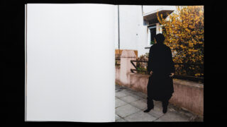 Ein Fotobuch, auf einer Seite aufgeschlagen, mit der Silhouette einer Person, die auf einem Bürgersteig neben einem gelb blühenden Busch und einem Wohngebäude steht; die gegenüberliegende Seite ist leer.