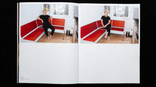 Eine Person in schwarzer Kleidung sitzt barfuß auf der Ecke einer rot-weißen Sitznische in einem spärlich dekorierten Raum, die in zweifacher Ausführung auf den aufgeschlagenen Seiten eines Buches abgebildet ist.