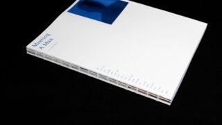 Auf einem schwarzen Untergrund liegt ein weißes Buch mit sichtbarer Fadenbindung und dem Titel „Meeting A Man“, auf dem Cover ist ein blau getöntes Foto zu sehen.