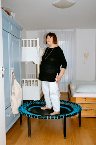 Eine ältere Frau steht auf einem Minitrampolin in einem Schlafzimmer mit Holzboden, einem Bett, einem Etagenbett und einem blauen Kleiderschrank.