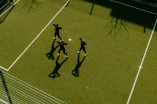 Drei schwarz gekleidete Personen stehen auf einem grünen Sportplatz im Freien und werfen im Sonnenlicht lange Schatten. Auf dem Boden sind weiße Begrenzungslinien und ein kleiner weißer Kreis zu sehen.