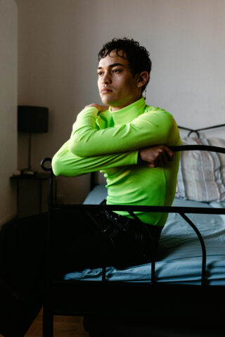 Eine Person in einem leuchtend grünen Langarmshirt sitzt in einem schlicht eingerichteten Zimmer auf der Bettkante und blickt mit neutralem Gesichtsausdruck zur Seite.