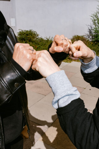 Im Freien berühren sich vier zu Fäusten geformte Hände, zwei Personen in dunklen Jacken sind teilweise zu sehen.