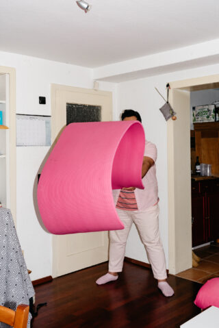 Eine Person in hellrosa Kleidung steht im Innenbereich und wirft oder hält eine große, zusammengerollte rosa Gymnastikmatte vor sich hin, sodass ihr Gesicht teilweise verdeckt ist.