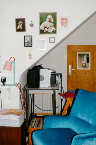 Ein blauer Sessel steht neben einem kleinen Tisch mit Audiogeräten unter einer mit gerahmten Hundefotos und Kunstwerken dekorierten Wand in einem gemütlichen Raum.