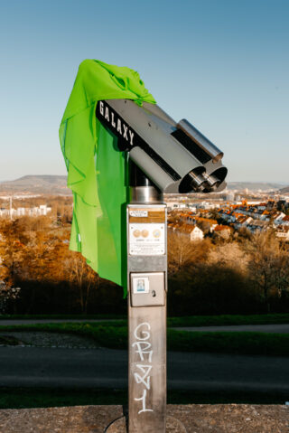 Ein öffentliches Beobachtungsteleskop mit der Aufschrift „GALAXY“, darüber ein neongrünes Tuch drapiert, mit Blick auf eine Stadtlandschaft und Hügel im Hintergrund.