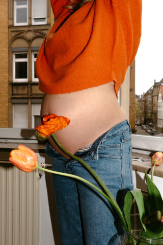 Eine Person trägt einen orangefarbenen Pullover und Bluejeans, wobei der Pullover hochgehoben ist und einen schwangeren Bauch freigibt. Sie steht auf einem Balkon neben einer Vase mit orangefarbenen Tulpen.