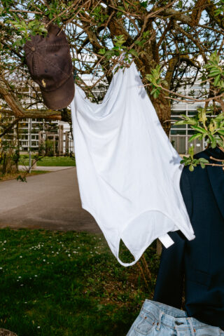 Ein weißes Tanktop und eine braune Baseballkappe hängen von den Zweigen eines Baumes, in der Nähe sind Jeans und eine dunkle Jacke teilweise zu sehen.