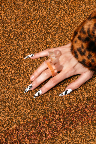 Eine Hand mit langen Nägeln im Kuhfellmuster und klobigen Ringen ruht auf einer strukturierten braunen Oberfläche. Die Person trägt einen Ärmel im Leopardenmuster.