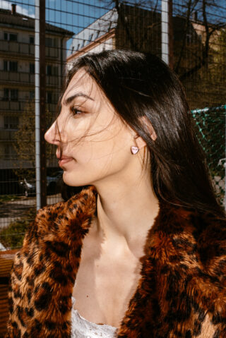 Eine Frau mit langem dunklem Haar, einem Mantel mit Leopardenmusterimitat und herzförmigen Ohrringen steht im Freien neben einem Maschendrahtzaun, im Hintergrund sind Gebäude und Bäume zu sehen.