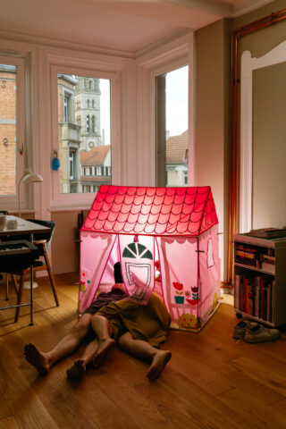 Eine Person liegt auf dem Boden und ihre Beine ragen aus einem kleinen rosa Spielhaus in einem hellen Wohnzimmer mit großen Fenstern.