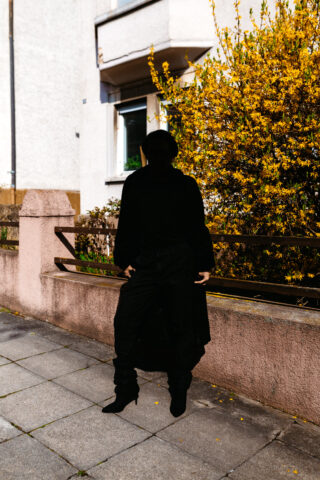 Eine ganz in Schwarz gekleidete Person steht auf einem Bürgersteig vor einem blühenden Busch und einem Gebäude, wobei ihr Gesicht und ihre Gesichtszüge durch Schatten verdeckt sind.