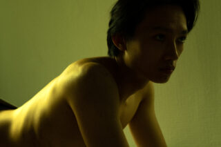 Eine oberkörperfreie Person mit kurzen dunklen Haaren ist im Seitenprofil zu sehen, wie sie sich unter schwachem, gelbgrünem Licht nach vorne beugt.