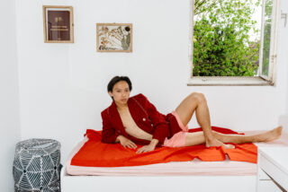 Eine Person in einem roten Blazer und rosa Shorts lümmelt auf einem Bett mit einem orangefarbenen Laken in einem weißen Raum mit offenem Fenster und gerahmter Kunst an der Wand.