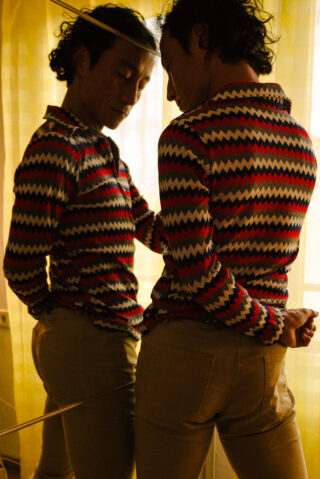 Eine Person in einem Hemd mit Zickzackmuster steht mit dem Rücken zur Kamera, blickt nach unten und ihr Spiegelbild ist in einem Spiegel neben ihr sichtbar.