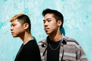 Zwei junge Männer stehen vor einer strukturierten türkisfarbenen Wand, einer blickt im Profil nach links und der andere nach vorne. Beide tragen Ohrringe und Freizeitkleidung.