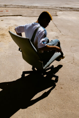 Eine Person sitzt im Freien auf einem rollenden Bürostuhl auf einer sonnenbeschienenen Betonfläche und wirft einen starken Schatten auf den Boden.