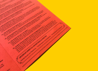 Auf einer gelben Fläche liegt ein rotes Dokument mit schwarzem deutschen Text, teilweise sichtbar, mit einem fettgedruckten Abschnitt mit der Aufschrift „INFORMATIONEN ZU UNTERLAGEN ENTFÄLLSCHREIBEN“.