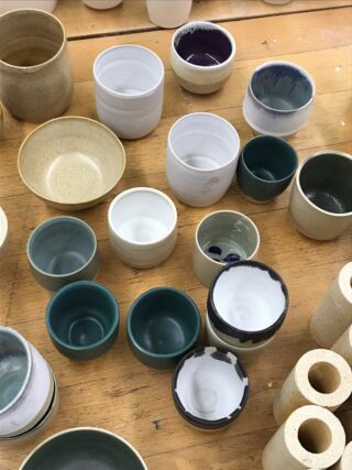 Verschiedene handgefertigte Tassen und Schüsseln aus Keramik in unterschiedlichen Größen und Farben sind auf einer Holztischplatte angeordnet.