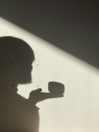 Der Schatten einer Person, die eine Tasse hält, wird auf eine sonnenbeschienene Wand geworfen, mit einem diagonalen Lichtstrahl in der oberen rechten Ecke.
