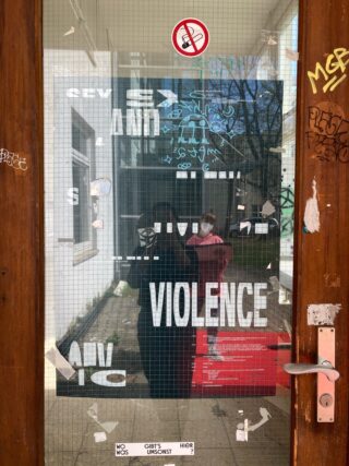 Eine Glastür mit Graffiti, Aufklebern und fettgedruckten, teilweise verdeckten weißen Texten mit den Aufschriften „SEX“, „GEWALT“ und anderen Wörtern. Im Glas spiegeln sich zwei Personen, eine davon trägt eine Gesichtsmaske.