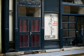 Eine dunkelblaue Ladenfront mit Glastür und Fenstern; ein Plakat mit einer Schlange und deutschem Text hängt daran, darunter „3 min ziehen lassen!“ in Rot.