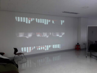 In einem minimalistisch eingerichteten Raum mit einem Stuhl, einem roten Hocker und verschiedenen Objekten in den Ecken wird eine Projektion abstrakter Bilder auf einer weißen Wand angezeigt.