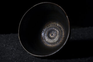 Eine dunkle, metallische Schale mit einem strukturierten, konzentrischen Kreismuster im Inneren, die auf einer schwarzen Oberfläche vor einem schwarzen Hintergrund ruht.