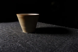 Eine einzelne Keramiktasse steht auf einer strukturierten dunklen Oberfläche, wird von links beleuchtet und hat einen Schatten nach rechts.