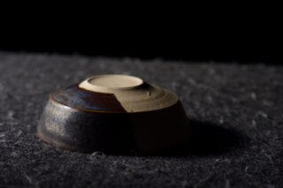 Eine Keramikschale, die verkehrt herum auf einer strukturierten dunklen Oberfläche steht, teilweise im Schatten, mit sichtbarem unglasiertem Ton auf der Unterseite.